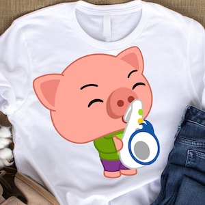 May include: White t-shirt featuring a cartoon pig playing a saxophone. The pig is pink with a green shirt and purple pants. The saxophone is white and blue. The shirt is paired with blue jeans.