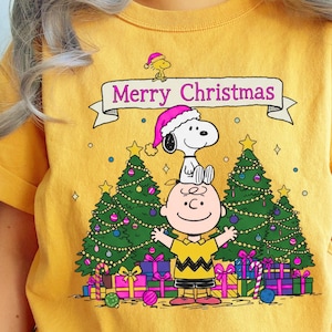 May include: Mustard yellow t-shirt featuring a graphic of Charlie Brown with Snoopy wearing Santa hats, and the text "Merry Christmas" on a banner. Christmas trees, presents, and candy canes complete the festive design.