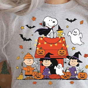 Op de afbeelding: Een grijze sweatshirt met een Halloween-thema. Het ontwerp toont Snoopy in een vampiercape, Charlie Brown, Lucy en andere Peanuts-personages verkleed voor Halloween, samen met pompoenen, vleermuizen en een spook.