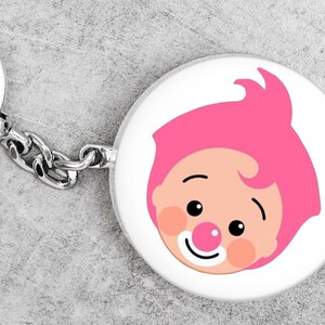 May include: A white, circular keyring featuring a cartoon clown's face. The clown has pink hair, rosy cheeks, a red nose, and a smiling mouth. The keyring has a silver chain and ring.