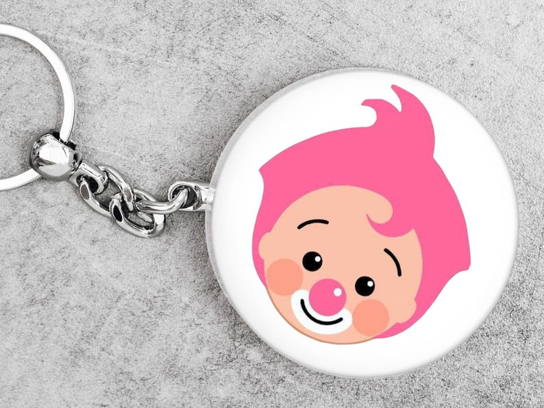 May include: A white, circular keyring featuring a cartoon clown's face. The clown has pink hair, rosy cheeks, a red nose, and a smiling mouth. The keyring has a silver chain and ring.