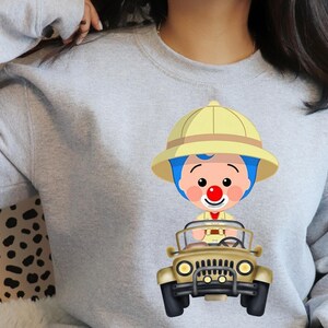May include: A pale grey sweatshirt with a cartoon image of a character wearing a safari hat and driving a beige jeep. The character has blue hair, a red nose, and a yellow safari hat.