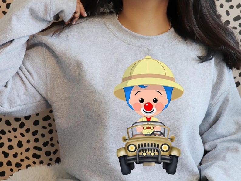 May include: A pale grey sweatshirt with a cartoon image of a character wearing a safari hat and driving a beige jeep. The character has blue hair, a red nose, and a yellow safari hat.