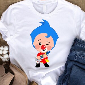 May include: White t-shirt with a cartoon character design. The character has blue hair, a red nose, and a red and yellow outfit, playing a guitar. The shirt has rolled-up sleeves, and a pair of blue jeans are visible.