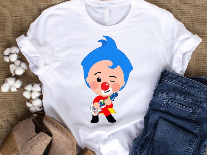May include: White t-shirt with a cartoon character design. The character has blue hair, a red nose, and a red and yellow outfit, playing a guitar. The shirt has rolled-up sleeves, and a pair of blue jeans are visible.