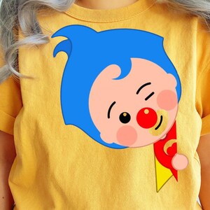 May include: Mustard yellow t-shirt with a cartoon character design. The character has blue hair, a red nose, and a pacifier. The character is winking and holding a red and yellow banner. The design is on the front.