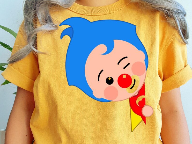 May include: Mustard yellow t-shirt with a cartoon character design. The character has blue hair, a red nose, and a pacifier. The character is winking and holding a red and yellow banner. The design is on the front.