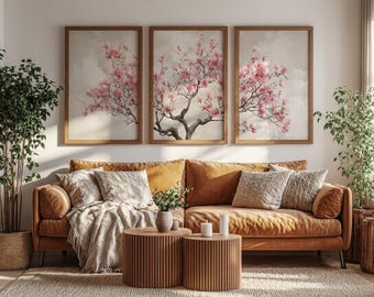Könnte beinhalten: Drei gerahmte Kunstdrucke mit Aquarellmalereien von rosa blühenden Bäumen. Die Kunstwerke hängen über einem braunen Sofa mit dekorativen Kissen und einer Decke. Zwei hölzerne Beistelltische und Pflanzen vervollständigen die Wohnzimmerdekoration.