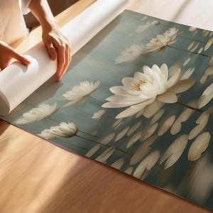 K&ouml;nnte beinhalten: Gerollter Kunstdruck mit einem Seerosen-Design. Der Druck, mit wei&szlig;en Seerosen und Seerosenbl&auml;ttern, wird auf einer Holzoberfl&auml;che ausgerollt. Das Kunstwerk hat eine Farbpalette aus T&uuml;rkis und Beige.