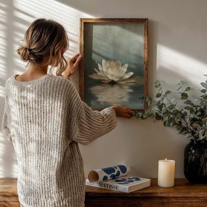 K&ouml;nnte beinhalten: Ein gerahmtes Kunstwerk mit einer wei&szlig;en Lotusblume, die auf Wasser schwimmt. Eine Frau h&auml;ngt das Kunstwerk an einer wei&szlig;en Wand auf. Eine Vase mit Gr&uuml;n, eine Kerze und ein Buch befinden sich auf einem Holztisch.