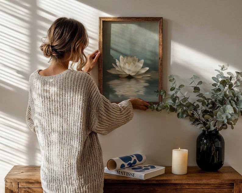 K&ouml;nnte beinhalten: Ein gerahmtes Kunstwerk mit einer wei&szlig;en Lotusblume, die auf Wasser schwimmt. Eine Frau h&auml;ngt das Kunstwerk an einer wei&szlig;en Wand auf. Eine Vase mit Gr&uuml;n, eine Kerze und ein Buch befinden sich auf einem Holztisch.