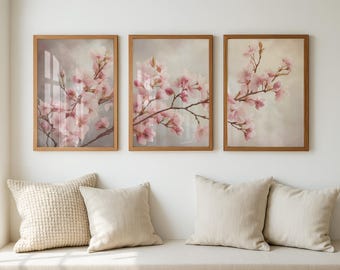 May include: Three framed art prints depicting pink blossom branches. The artwork has a soft, muted colour palette with a light background. The frames are a warm, natural wood tone. The prints are displayed above a bench with beige cushions.