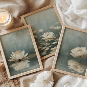 K&ouml;nnte beinhalten: Drei gerahmte Kunstdrucke mit wei&szlig;en Lotusbl&uuml;ten auf Wasser, in einem weichen blau-gr&uuml;nen Aquarellstil. Die Rahmen sind aus hellem Holz. Eine brennende Kerze und Trockenblumen sind ebenfalls vorhanden.