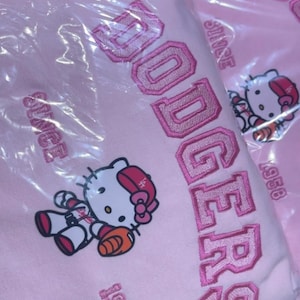 K&ouml;nnte beinhalten: Hellrosa Kapuzenpullover mit dem Wort "Dodgers" in rosa gestickt. Das Design umfasst eine Hello Kitty-Grafik in Baseball-Uniform und den Text "Since 1958".