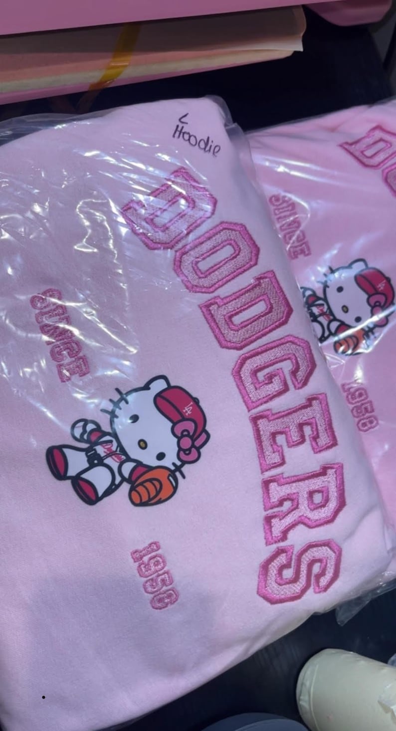 K&ouml;nnte beinhalten: Hellrosa Kapuzenpullover mit dem Wort "Dodgers" in rosa gestickt. Das Design umfasst eine Hello Kitty-Grafik in Baseball-Uniform und den Text "Since 1958".