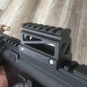 Op de afbeelding: Zwarte metalen geweerkijkerbevestiging met een Picatinny-rail. De bevestiging is bevestigd aan een zwarte geweerkast met zilveren schroeven. De achtergrond is een lichtbruin houten oppervlak.