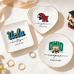 May include: Three white ceramic ring dishes with gold rims. One square dish features "Ucla" in blue and black, with "kaittyn" and "CLASS OF 2026". A heart-shaped dish has a red eagle logo and "hayden, CLASS OF 2026". A round dish displays "OHIO" and "cooper, CLASS OF 2026".