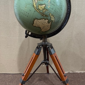 Może przedstawiać: Globus w stylu vintage w odcieniach turkusu i beżu, zamontowany na drewnianym statywie. Globus przedstawia szczegóły geograficzne i jest podtrzymywany przez ciemną metalową ramę. Drewniane nogi stojaka są w ciepłym brązowym kolorze.