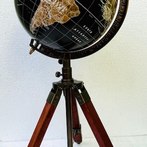 Może przedstawiać: Dekoracyjny globus z czarną powierzchnią i beżowymi kontynentami, zamontowany na statywie z ciemnego drewna i metalu. Globus posiada szczegółowe oznaczenia geograficzne. Statyw ma nogi z ciemnoczerwonego drewna.