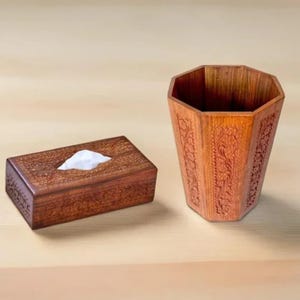 Könnte beinhalten: Eine Holz-Taschentuchbox und ein passender Abfalleimer. Beide Artikel sind warmbraun und weisen aufwendige Blumenschnitzereien auf. Die Taschentuchbox hat eine rechteckige Form. Der Abfalleimer hat eine achteckige Form.