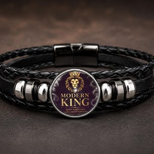 May include: A black braided leather bracelet featuring a central circular charm with a gold lion head graphic and the text MODERN KING. The bracelet includes silver-toned metal accents and a black magnetic clasp closure on a dark background.