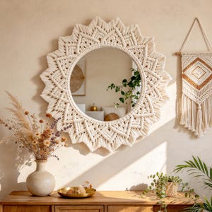 May include: A round wall mirror featuring an intricate off-white macrame frame with a sunburst design. The knotted cotton cord creates a geometric pattern around the circular glass surface, providing a textured border that radiates outward.