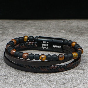 Peut inclure: Un bracelet multicouche composé de cuir marron et de brins perlés. Le bracelet comprend des perles noires, des perles œil de tigre et un fermoir noir gravé de "I am so proud of you!" et un symbole de cœur suivi de "Mom".