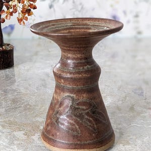 May include: A brown ceramic candle holder with a wide, shallow top and a flared base. The body features a textured surface with leaf-like patterns. The candle holder is approximately 15 cm tall.