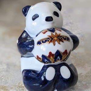 May include: A ceramic panda ornament in white and navy blue, holding a decorative egg. The egg has a floral design with gold, red, and blue accents. The panda's ears and limbs are navy blue.