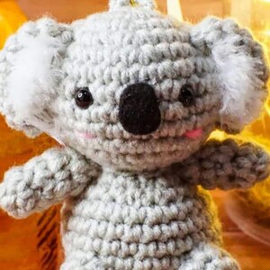 May include: A handmade crochet koala bear in shades of grey, with a black nose, small black eyes, and pink accents on the cheeks. The koala has fluffy white ears and a loop for hanging. A cute, soft toy.