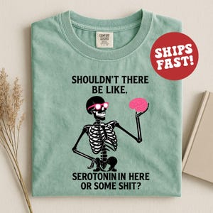 May include: A light green t-shirt featuring a black skeleton wearing pink sunglasses and holding a pink brain. The text on the shirt reads "SHOULDN'T THERE BE LIKE, SEROTONIN IN HERE OR SOME SHIT?" A red circle with "SHIPS FAST!" is in the upper right.
