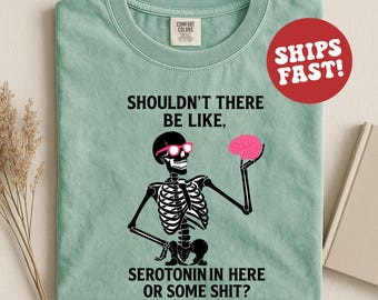 Può includere: Maglietta verde chiaro con uno scheletro nero con occhiali da sole rosa e un cervello rosa. Il testo sulla maglietta recita: "SHOULDN'T THERE BE LIKE, SEROTONIN IN HERE OR SOME SHIT?" Un cerchio rosso con "SHIPS FAST!" è in alto a destra.
