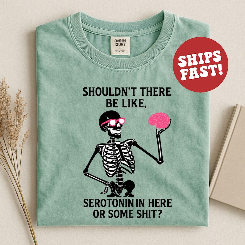 May include: A light green t-shirt featuring a black skeleton wearing pink sunglasses and holding a pink brain. The text on the shirt reads "SHOULDN'T THERE BE LIKE, SEROTONIN IN HERE OR SOME SHIT?" A red circle with "SHIPS FAST!" is in the upper right.
