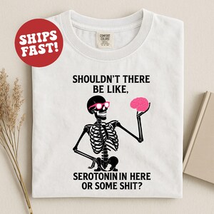 May include: White t-shirt with a black skeleton graphic wearing pink glasses and holding a pink brain. The text reads "SHOULDN'T THERE BE LIKE, SEROTONIN IN HERE OR SOME SHIT?" A red circle with "SHIPS FAST!" is in the upper left.