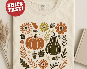 May include: Cream-colored t-shirt featuring a fall-themed design. The design includes two pumpkins, flowers, leaves, and acorns in shades of brown, green, and orange. The shirt has a crew neck and short sleeves. The text "COMFORT COLORS" is visible on the tag.