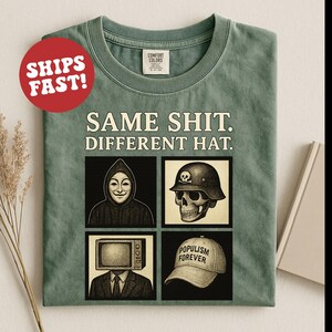Könnte beinhalten: Salbeigrünes T-Shirt mit dem Text "SAME SHIT. DIFFERENT HAT." und vier quadratischen Illustrationen. Die Illustrationen zeigen eine Guy-Fawkes-Maske, einen Schädel in einem Helm, eine Person mit einem Fernseh-Kopf und einen "POPULISM FOREVER" Hut.