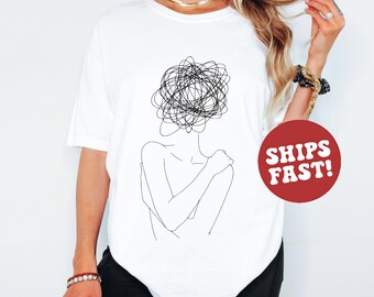 May include: White t-shirt featuring a minimalist line art design of a person with a scribbled head. The black graphic is centered on the shirt. The shirt is worn by a person. The image also includes a red circle with the words "SHIPS FAST!"