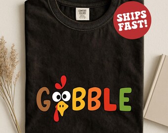 May include: Black t-shirt with the word "GOBBLE" in colorful letters, featuring a turkey face design. The shirt is made of a soft material and is perfect for Thanksgiving.