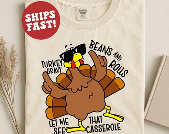 Puede incluir: Camiseta color crema con un pavo de dibujos animados con gafas de sol. El pavo tiene plumas marrones y patas amarillas, con el texto "TURKEY GRAVY, BEANS AND ROLLS, LET ME SEE THAT CASSEROLE". Un círculo rojo con "SHIPS FAST!" está en la parte superior izquierda.