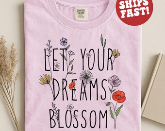 Puede incluir: Camiseta rosa claro con la frase "LET YOUR DREAMS BLOSSOM" en negro, rodeada de ilustraciones florales coloridas. Un círculo rojo con la frase "SHIPS FAST!" está en la esquina superior derecha.