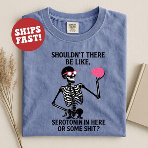 May include: A blue t-shirt featuring a skeleton wearing pink sunglasses and holding a pink brain. The text on the shirt reads, "SHOULDN'T THERE BE LIKE, SEROTONIN IN HERE OR SOME SHIT?" A red circle with "SHIPS FAST!" is in the upper left.