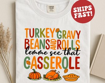 Puede incluir: Camiseta blanca con un gráfico colorido con temática de Acción de Gracias. El texto dice "TURKEY GRAVY BEANS AND ROLLS lemme see that CASSEROLE" con imágenes de calabaza, pavo y tarta. La camiseta tiene una etiqueta Comfort Colors.