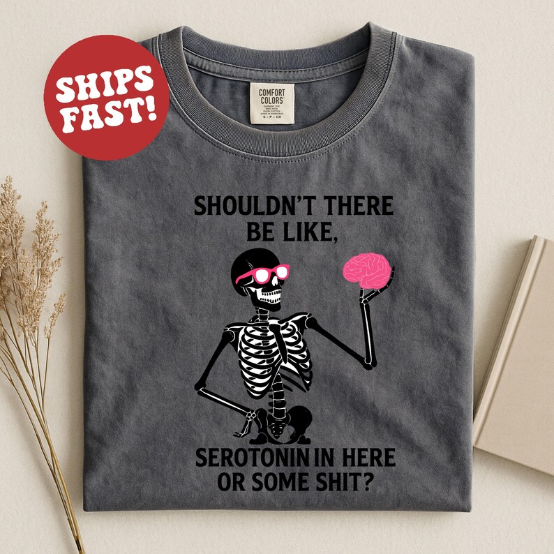 May include: Dark gray t-shirt with a skeleton graphic wearing pink sunglasses and holding a pink brain. The text on the shirt reads "SHOULDN'T THERE BE LIKE, SEROTONIN IN HERE OR SOME SHIT?"