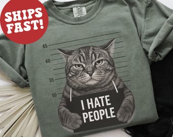 May include: Sage green t-shirt featuring a grumpy cat mugshot with a sign that reads "I HATE PEOPLE." The cat has gray and black stripes. The shirt has a vintage, washed look.