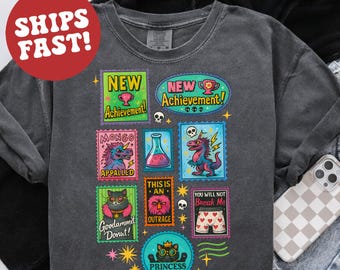 May include: Dark gray sweatshirt with colorful cartoon stamp designs. Stamps read "NEW Achievement!", "MONGO Appalled", "THIS IS AN OUTRAGE", "Goodammit Donut!", "PRINCESS DONUT", and "YOU WILL NOT Break Me". A red circle with "SHIPS FAST!" is in the upper left.
