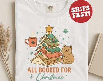 Op de afbeelding: Wit T-shirt met een kerstthema. Het ontwerp toont een stapel boeken in de vorm van een kerstboom, een kat, een mok en de tekst "ALL BOOKED FOR Christmas". Een rode cirkel met "SHIPS FAST!" is ook zichtbaar.
