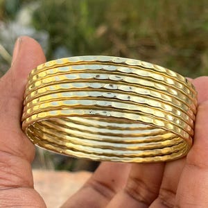 May include: A stack of eight gold-colored bangles with a hammered texture. The bangles are round and appear to be made of metal. The background is blurred, with green and brown tones.