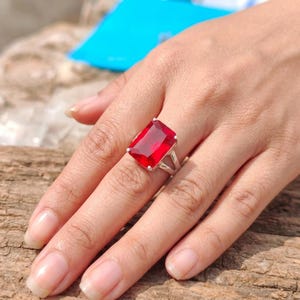 Può includere: Un anello in argento con una grande pietra preziosa rossa rettangolare sfaccettata, fissata con una montatura a quattro griffe. La pietra è di colore rosso intenso e trasparente, cattura la luce con i suoi bordi netti e la superficie lucida.