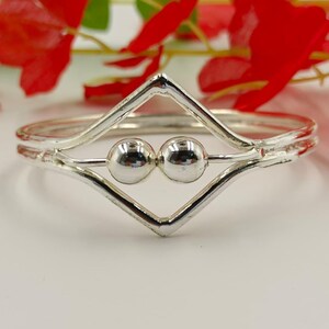 May include: A silver bracelet with a geometric design. The bracelet features a diamond-shaped center with two small, round silver beads. The band is a simple, thin silver band. The background includes red flowers and green leaves.