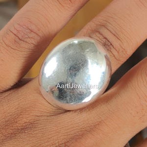 May include: A polished silver ring featuring a large, smooth, oval-shaped centerpiece with a reflective surface. The metal has a bright, metallic sheen and a slightly irregular, organic contour. The text AartiJewellers is printed on the center.